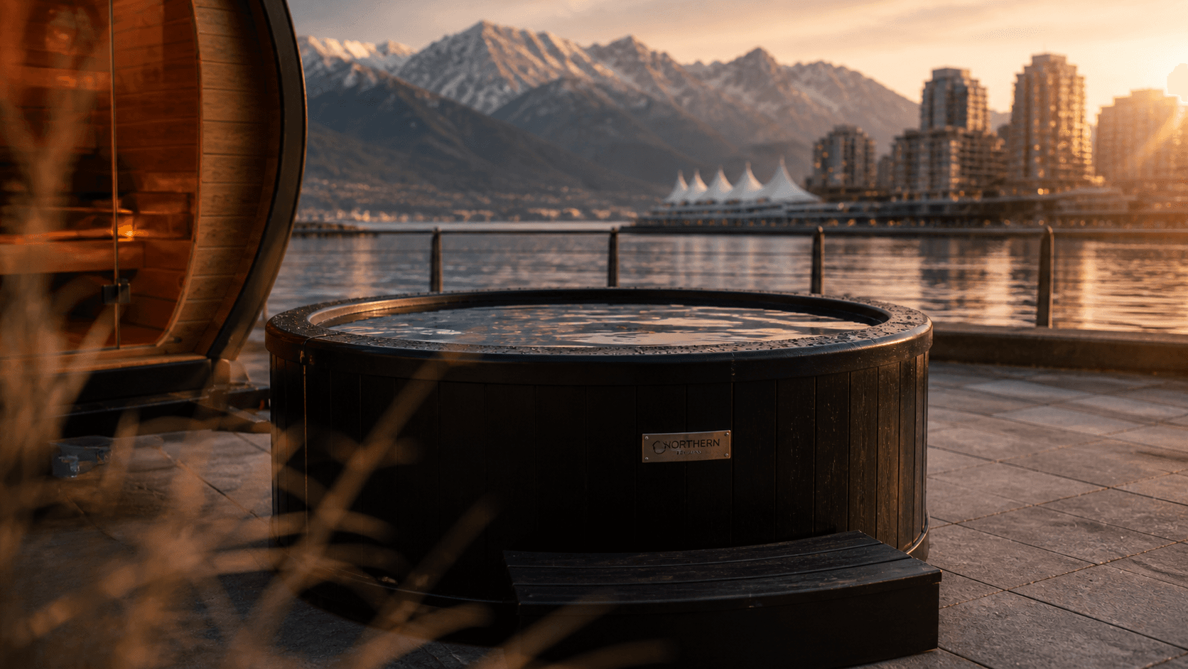 Outdoor hot tub on a stone patio overlooking a city skyline and snow-capped mountains at sunset.